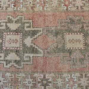 Puede incluir: Una alfombra vintage con un esquema de color rosa y crema descolorido. La alfombra presenta un patr&oacute;n geom&eacute;trico repetitivo con un dise&ntilde;o central de dos grandes motivos en forma de estrella de color verde oscuro.