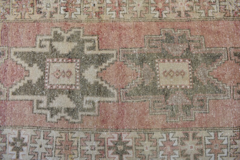 Puede incluir: Una alfombra vintage con un esquema de color rosa y crema descolorido. La alfombra presenta un patr&oacute;n geom&eacute;trico repetitivo con un dise&ntilde;o central de dos grandes motivos en forma de estrella de color verde oscuro.