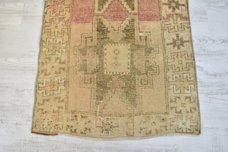 Puede incluir: Una alfombra turca vintage de corredor con fondo beige y intrincados patrones geom&eacute;tricos en tonos marrones y rosados. La alfombra tiene un borde con flecos y un aspecto desgastado y envejecido.
