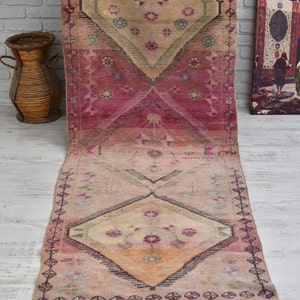 K&ouml;nnte beinhalten: Ein langer, rechteckiger rosa-beiger Teppich mit einem Blumenmuster. Der Teppich hat eine Diamantform in der Mitte mit einem Blumendesign. Der Teppich hat an beiden Enden Fransen.