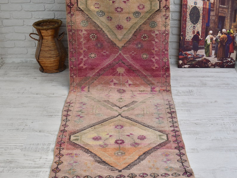 K&ouml;nnte beinhalten: Ein langer, rechteckiger rosa-beiger Teppich mit einem Blumenmuster. Der Teppich hat eine Diamantform in der Mitte mit einem Blumendesign. Der Teppich hat an beiden Enden Fransen.