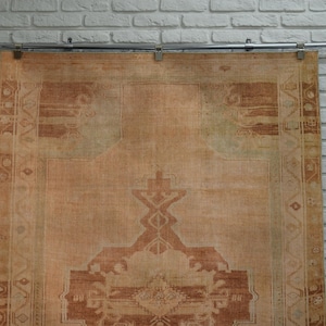 May include: A large, rectangular, vintage rug with a faded, distressed appearance. The rug features a central medallion design in shades of brown and beige, with intricate border patterns. The rug is hanging against a white brick wall.