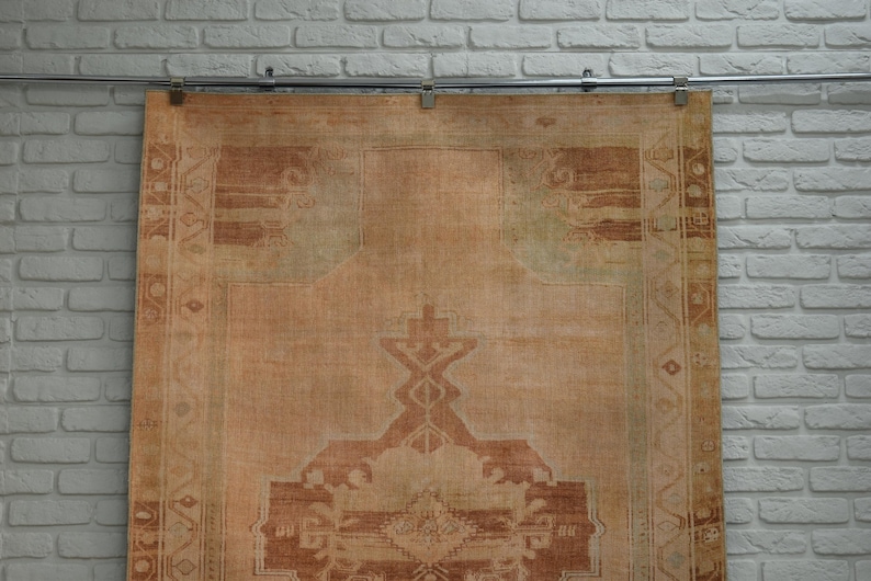 May include: A large, rectangular, vintage rug with a faded, distressed appearance. The rug features a central medallion design in shades of brown and beige, with intricate border patterns. The rug is hanging against a white brick wall.
