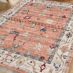 May include: A peach-colored rug with a floral and geometric pattern. The rug has a cream-colored border with a blue and gray inner border.