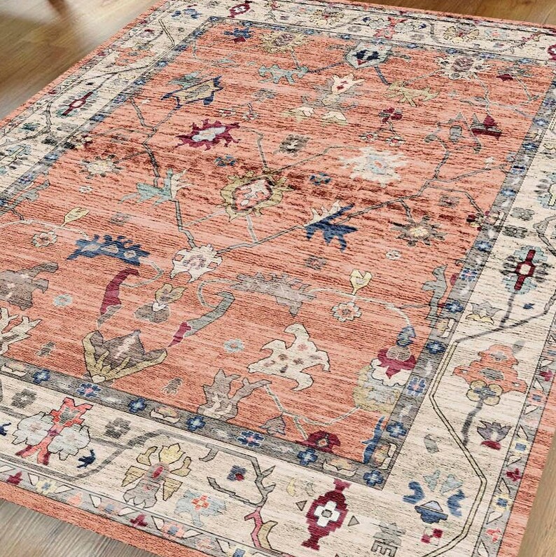 May include: A peach-colored rug with a floral and geometric pattern. The rug has a cream-colored border with a blue and gray inner border.
