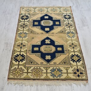 May include: A beige rug with a traditional geometric pattern in shades of blue, yellow, and green. The rug has a fringe border.