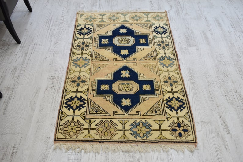 May include: A beige rug with a traditional geometric pattern in shades of blue, yellow, and green. The rug has a fringe border.