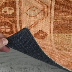 May include: Close-up of a section of a rug with a geometric pattern in shades of orange and brown. The rug's underside is a dark blue-gray color. The rug is being held up, revealing the underside.