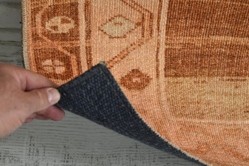 May include: Close-up of a section of a rug with a geometric pattern in shades of orange and brown. The rug's underside is a dark blue-gray color. The rug is being held up, revealing the underside.