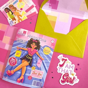 May include: A pink and yellow themed flat lay featuring a magazine, coupons, an envelope, and a sticker. The magazine cover reads "Mini Magazine" with a cartoon illustration. The sticker says "I AM THAT Girl".