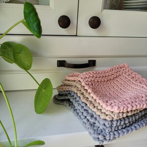 May include: A stack of four knitted dishcloths in pink, beige, and grey. The dishcloths are folded and stacked on a white countertop.