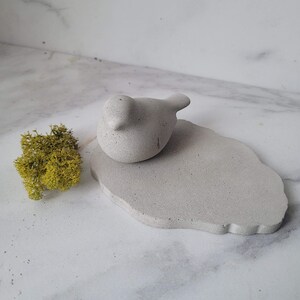 May include: A small, gray concrete bird figurine perched on a larger, gray concrete base with an irregular shape. The base has a rough, textured surface.