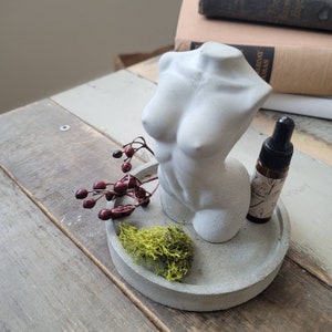Peut inclure: Une sculpture en béton d'un torse avec un petit plateau en béton, une brindille de baies rouges et une petite bouteille d'huile essentielle. Le plateau est sur une surface en bois.