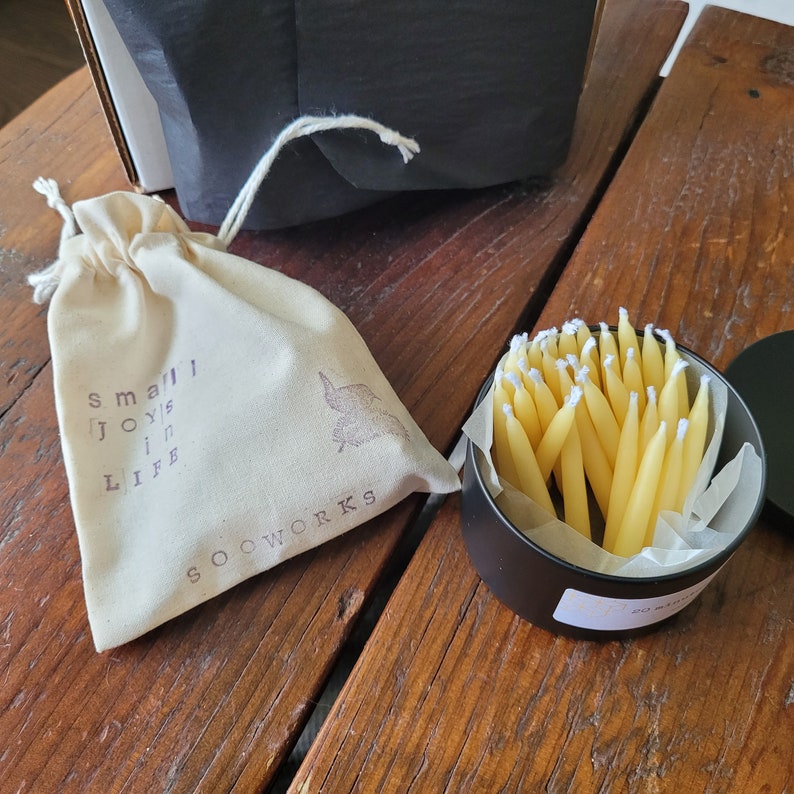May include: A small white fabric bag with the text "small joys in life" and the word "SOOWORKS" printed on it. The bag is next to a black metal container filled with yellow birthday candles.