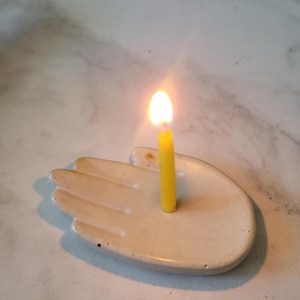 May include: A white ceramic hand-shaped candle holder with a yellow candle burning inside.