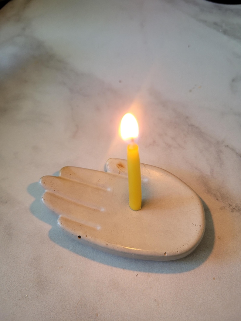May include: A white ceramic hand-shaped candle holder with a yellow candle burning inside.