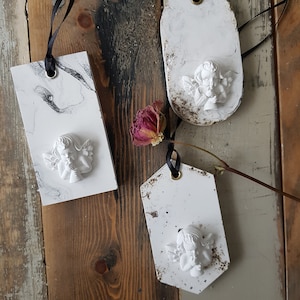 May include: Three decorative tags with white cherub angel designs. One tag has a black and white marble pattern, while the others have a distressed, aged appearance. Each tag is strung with a black ribbon and has a gold eyelet.
