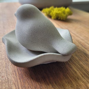 May include: A gray ceramic bird figurine perched on a matching leaf-shaped dish. The bird has a textured surface and a rounded body. The dish has a wavy edge. The background includes a wooden surface and a blurred gray pot.