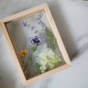May include: A wooden frame containing a pressed flower arrangement. The flowers are purple, yellow, and white. The frame is a light wood colour.