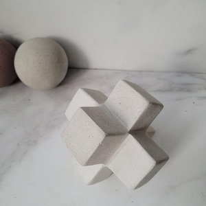 May include: A white concrete geometric sculpture in the shape of a cross. The sculpture has a rough, textured surface.