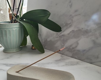 May include: A beige, rectangular incense holder with a single hole, holding a lit incense stick. The holder sits on a white marble surface, with a light blue ceramic pot and an orchid plant in the background.