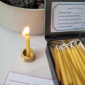 May include: A set of 10 small, yellow beeswax candles in a black metal tin. The candles are arranged in a row and are ready to be used for meditation or mindfulness practices. The tin has a label that reads "Handle With Care" and includes instructions for burning the candles safely.