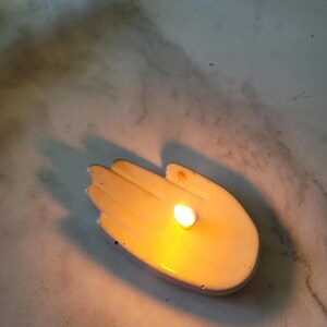 May include: A white ceramic hand-shaped candle holder with a lit candle inside.