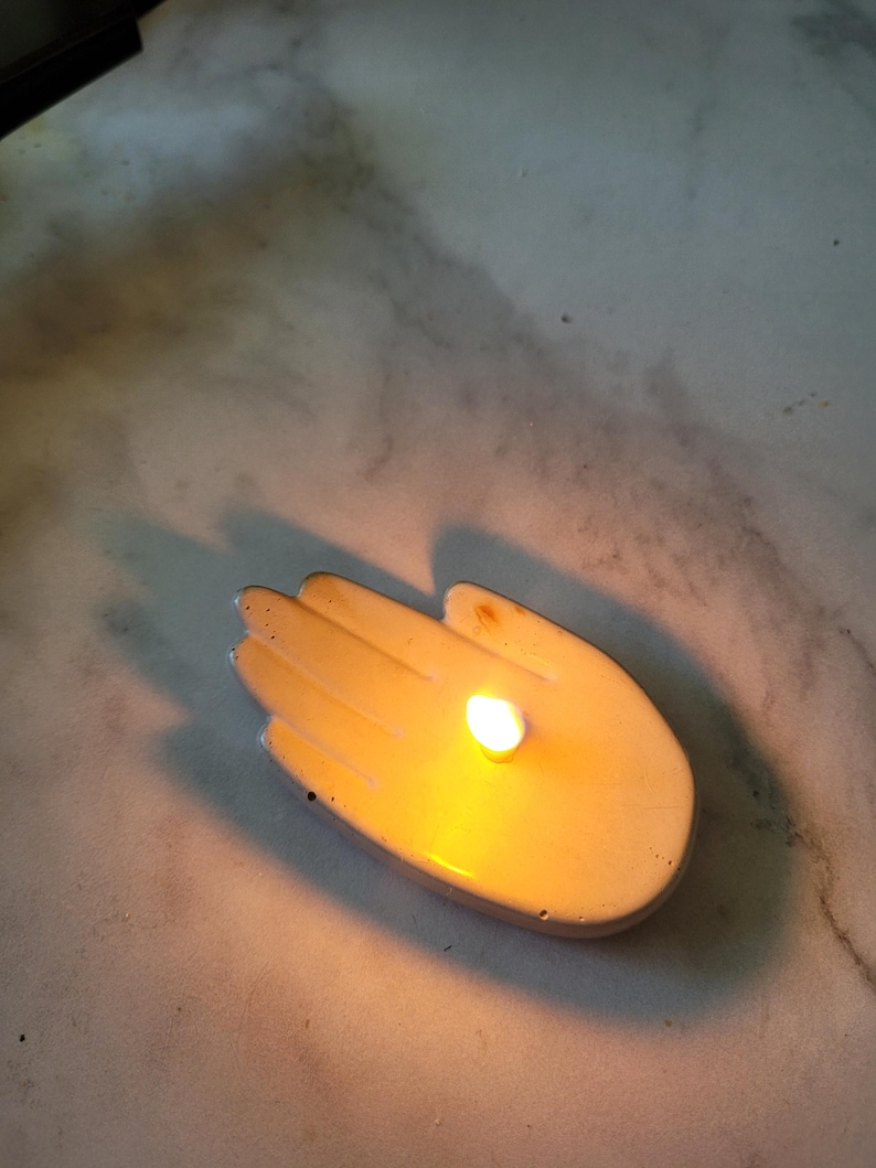 May include: A white ceramic hand-shaped candle holder with a lit candle inside.