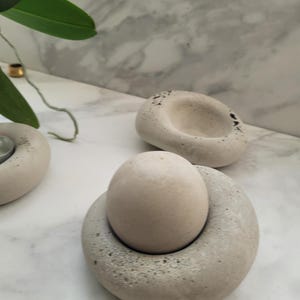 May include: A set of three light grey, stone-like candle holders. One holds a small, egg-shaped object. The other two have a circular design with a small indentation. The objects are on a white marble surface.