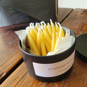 May include: A black metal tin filled with yellow birthday candles. The tin has a white label with the text "20 minutes beeswax 100% pure candle"