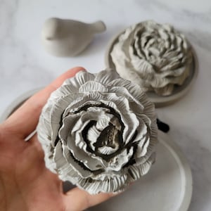 May include: Two grey concrete flower sculptures. One is a single flower with multiple layers of petals. The other is a cluster of flowers.