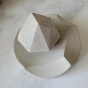 Peut inclure: Un ensemble de deux sculptures géométriques en béton de couleur gris clair. La plus grande sculpture est un plat plat de forme ovale avec un bord légèrement surélevé. La plus petite sculpture est une pyramide géométrique qui repose sur le plat.