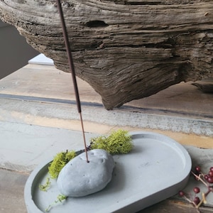 May include: A grey concrete incense holder with a small grey stone in the centre and green moss around the stone. A brown incense stick is sticking straight up from the stone.