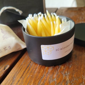 May include: A black cylindrical container with a white label that says "20 minutes beauty". The container is filled with a bundle of yellow beeswax candles.