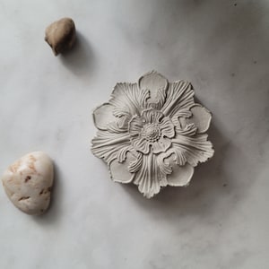 May include: A white ceramic flower with intricate details, resembling a stylised floral design. The flower is on a white background.