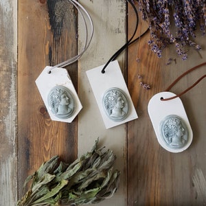 May include: Three white rectangular tags with a gray cameo image of a woman with flowers in her hair. The tags are hanging from white and black cords.