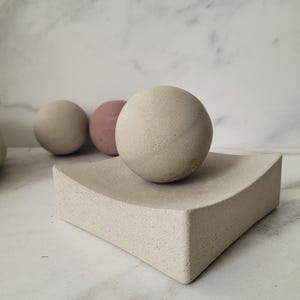 May include: Three smooth, round, concrete spheres, one of which is resting on a curved, square, concrete base. The spheres are in shades of grey and pink.