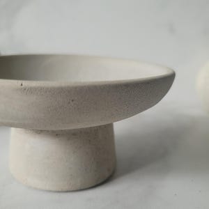Peut inclure: Un bol en béton blanc sur un piédestal rond. Le bol est lisse et a une surface légèrement texturée.