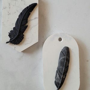 May include: Two white ceramic ornaments with black feather designs. The ornaments are shaped like a hexagon and an oval.