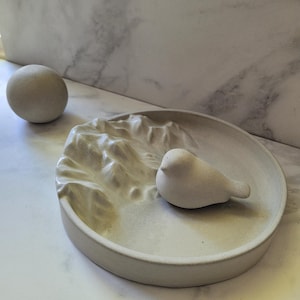 May include: A light grey decorative tray with a mountain range design and a small bird figurine. A grey sphere sits to the left of the tray. The objects are made of a stone-like material.