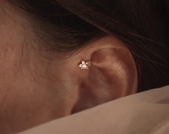 Puede incluir: Un pendiente de cartílago dorado con forma de pequeña flor de cinco pétalos. El centro de la flor está engastado con una piedra preciosa transparente y brillante, rodeada por cuatro piedras claras más pequeñas en cada pétalo.