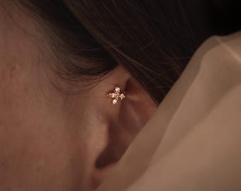 Peut inclure: Une boucle d'oreille en métal doré composée de quatre petites pierres transparentes taille marquise disposées en forme de fleur, fixée sur le cartilage supérieur de l'oreille, captant la lumière avec un éclat discret sur la peau.