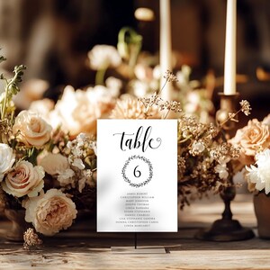 May include: A white table number card with the word "table" in elegant script and the number "6" inside a wreath. Below the number are guest names. The card is displayed on a metal stand, surrounded by floral arrangements and candles.