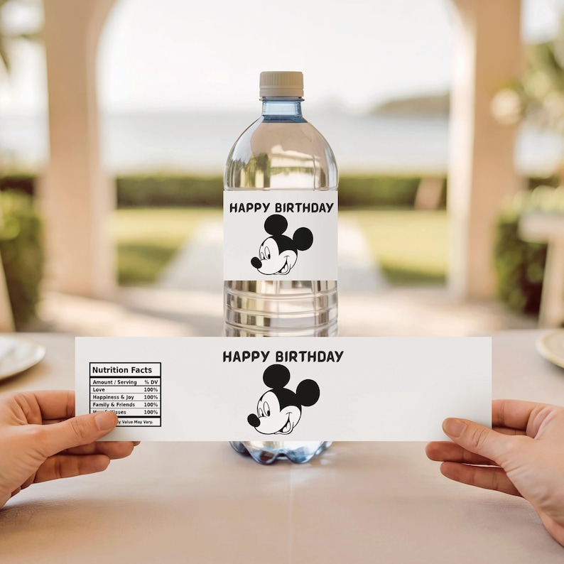 May include: A clear plastic water bottle with a white label that reads "HAPPY BIRTHDAY" and features a black and white Mickey Mouse graphic. A second label with the same design is held below the bottle. The background is blurred.