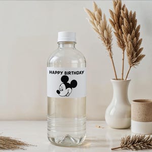 May include: A clear plastic water bottle with a white cap and a label that reads "HAPPY BIRTHDAY" with a Mickey Mouse graphic. The bottle is next to a white vase with dried plants and a small ceramic cup.