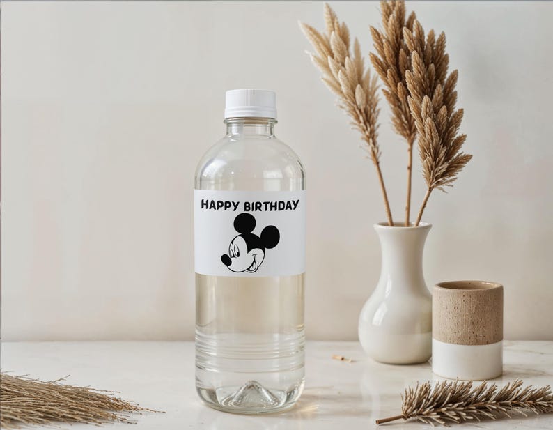 May include: A clear plastic water bottle with a white cap and a label that reads "HAPPY BIRTHDAY" with a Mickey Mouse graphic. The bottle is next to a white vase with dried plants and a small ceramic cup.