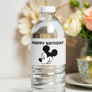 May include: Clear plastic water bottle with a white cap. The label features the text "HAPPY BIRTHDAY" above a black and white illustration of Mickey Mouse. The bottle is filled with clear liquid. Flowers and a teacup are in the background.