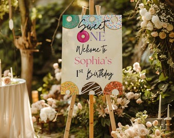 Könnte beinhalten: Eine Holzstaffelei zeigt ein Schild mit den Worten "Sweet One" und "Welcome to Sophia's 1st Birthday". Das Schild ist mit Donut-Illustrationen verziert. Der Hintergrund ist eine verschwommene Außenszene mit Blumen und Blättern.