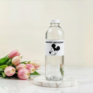 May include: A clear glass bottle with a white cap and a label that reads "HAPPY BIRTHDAY" with a Mickey Mouse graphic. The bottle sits on a marble coaster next to a bouquet of pink tulips.
