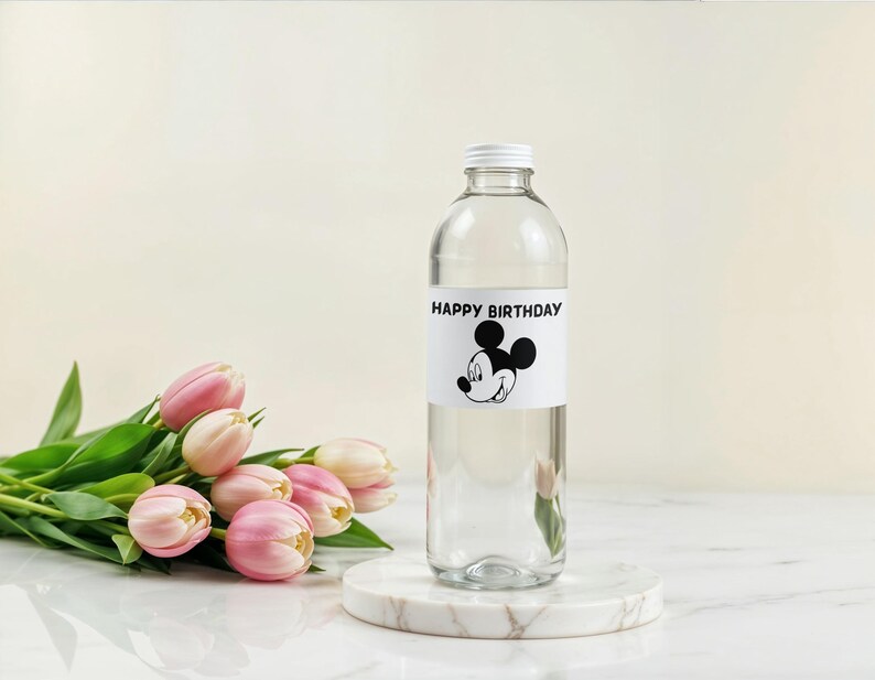 May include: A clear glass bottle with a white cap and a label that reads "HAPPY BIRTHDAY" with a Mickey Mouse graphic. The bottle sits on a marble coaster next to a bouquet of pink tulips.